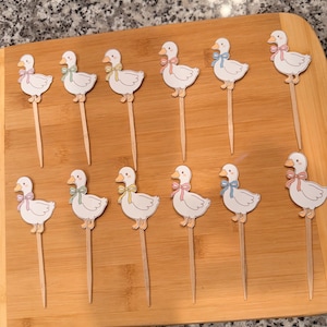 Può includere: Dodici decorazioni per cupcake a forma di oca bianca con fiocchi pastello rosa, verdi e blu. Ogni oca è attaccata a uno stecchino di legno, perfette per decorare i prodotti da forno. Le decorazioni sono disposte su un tagliere di legno.