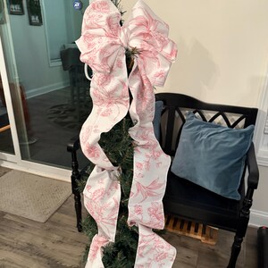May include: A small Christmas tree decorated with a large bow. The bow is white with a pink floral pattern. The tree is in a copper-colored pot. The background includes a black bench with a blue pillow.