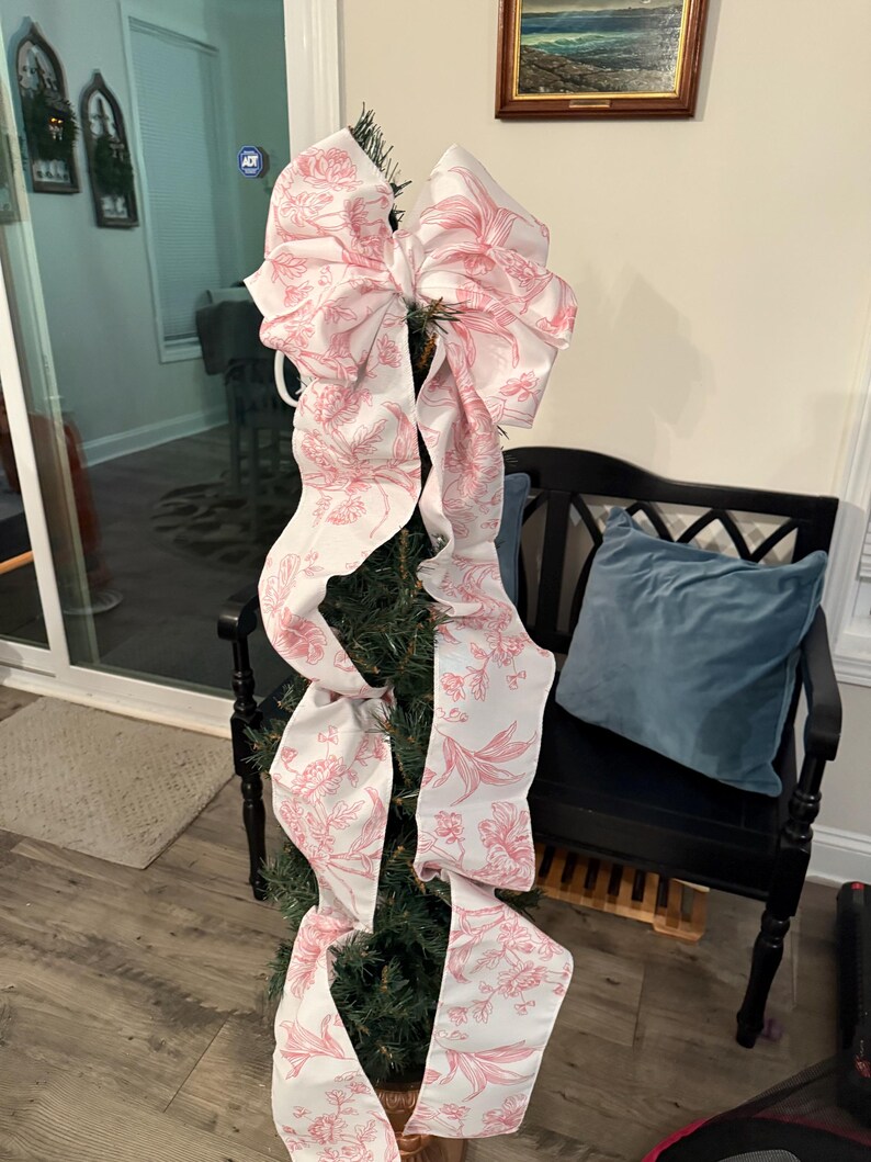 May include: A small Christmas tree decorated with a large bow. The bow is white with a pink floral pattern. The tree is in a copper-colored pot. The background includes a black bench with a blue pillow.