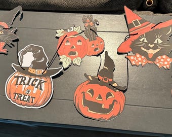 May include: A Halloween-themed garland featuring six hanging cutouts: a black cat with a red bow, an owl on a red crescent moon, a pumpkin with the text TRICK OR TREAT, a pumpkin with a black cat, a cat in a witch hat, and an owl on a pumpkin.