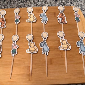 Könnte beinhalten: Ein Set aus zwölf dekorativen Lebensmittelspießen mit Illustrationen von Winnie Puuh-Figuren, darunter Tigger, Ferkel, Puuh und I-Aah. Jede Figur hält einen runden Luftballon und ist an einem dünnen Holzstab befestigt.