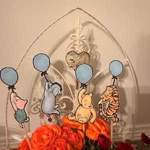 May include: Four paper cutouts of cartoon characters Piglet, Eeyore, Winnie the Pooh, and Tigger, each holding a light blue balloon. The characters are attached to wooden sticks and arranged among orange and red artificial flowers.