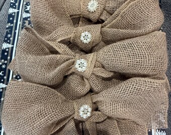 May include: Four large, rustic burlap bows with sparkling rhinestone centers. The bows are a warm tan color and are arranged vertically. The background features a festive pattern with white snowflakes and trees.