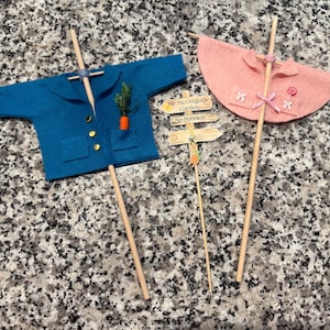 以下が含まれることがあります： 青いジャケットとピンクのマントの2つのフェルトトッパー。ポケットにはニンジンが入っています。「Mr. McGregor's Garden, No Bunnies!」と書かれた看板。すべて木製の棒に取り付けられており、ケーキのデコレーションや工芸に最適です。