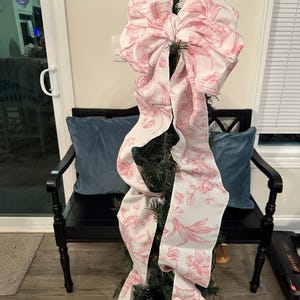 May include: A small Christmas tree decorated with a large white bow featuring a pink floral pattern. The bow's long ribbons cascade down the tree. The tree is placed in a black bench with blue pillows.