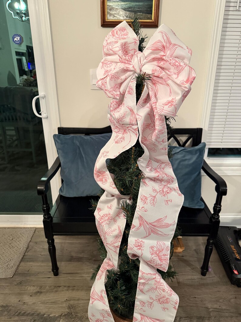 May include: A small Christmas tree decorated with a large white bow featuring a pink floral pattern. The bow's long ribbons cascade down the tree. The tree is placed in a black bench with blue pillows.
