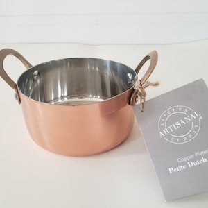 Könnte beinhalten: Ein kupferbeschichteter Mini-Dutch-Oven mit Edelstahl-Innenraum und bronzefarbenen Griffen. Das Etikett lautet "Kitchen Artisanal Supply Copper Plated Petite Dutch Oven".