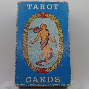 Puede incluir: Una caja de cartas del tarot vintage con un fondo azul y las palabras "TAROT CARDS" en blanco. La caja presenta una ilustraci&oacute;n ovalada de una figura desnuda sosteniendo velas, rodeada por una corona dorada y cintas rojas.