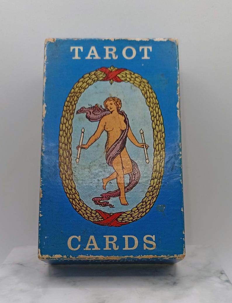 Puede incluir: Una caja de cartas del tarot vintage con un fondo azul y las palabras "TAROT CARDS" en blanco. La caja presenta una ilustraci&oacute;n ovalada de una figura desnuda sosteniendo velas, rodeada por una corona dorada y cintas rojas.