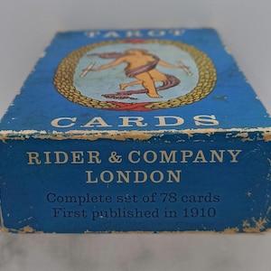 Puede incluir: Caja de cartas de tarot azul vintage con letras doradas y una ilustraci&oacute;n de una figura. La caja dice "TAROT CARDS" y "RIDER & COMPANY LONDON". La caja es un juego completo de 78 cartas, publicado por primera vez en 1910.