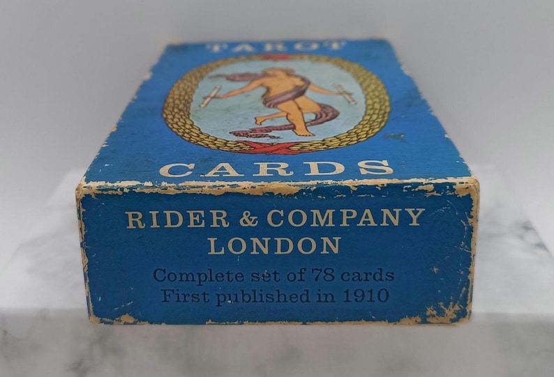 Puede incluir: Caja de cartas de tarot azul vintage con letras doradas y una ilustraci&oacute;n de una figura. La caja dice "TAROT CARDS" y "RIDER & COMPANY LONDON". La caja es un juego completo de 78 cartas, publicado por primera vez en 1910.
