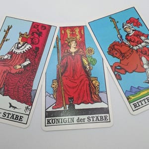 Op de afbeelding: Drie tarotkaarten met illustraties van figuren in rode gewaden en harnassen tegen een blauwe achtergrond. De kaarten tonen de Koning, Koningin en Ridder van Staven, met Duitse tekst onderaan.