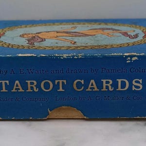 Puede incluir: Una caja de cartas de tarot azul vintage con las palabras "TAROT CARDS" en letras blancas grandes. La caja presenta una ilustraci&oacute;n de dos figuras dentro de un marco ovalado. Tambi&eacute;n es visible el texto "Conceived by A. E. Waite and drawn by Pamela Colman Smith".