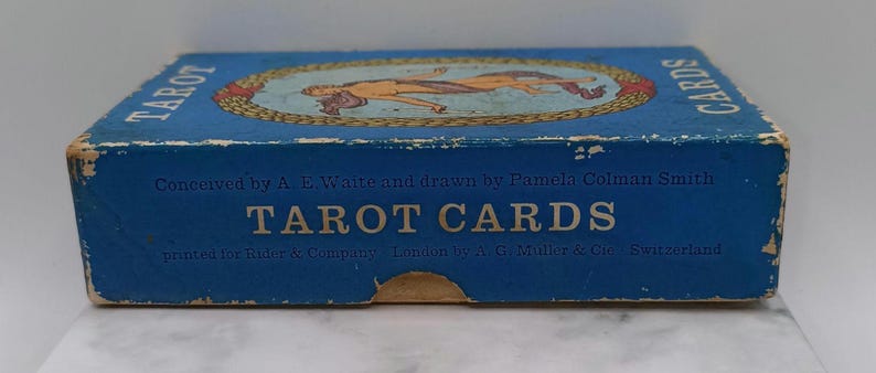 Puede incluir: Una caja de cartas de tarot azul vintage con las palabras "TAROT CARDS" en letras blancas grandes. La caja presenta una ilustraci&oacute;n de dos figuras dentro de un marco ovalado. Tambi&eacute;n es visible el texto "Conceived by A. E. Waite and drawn by Pamela Colman Smith".
