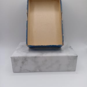 Puede incluir: Una caja rectangular abierta con borde azul e interior beige se encuentra sobre un bloque de m&aacute;rmol blanco. La caja parece ser vintage, con signos de desgaste. El bloque de m&aacute;rmol tiene un patr&oacute;n veteado gris.