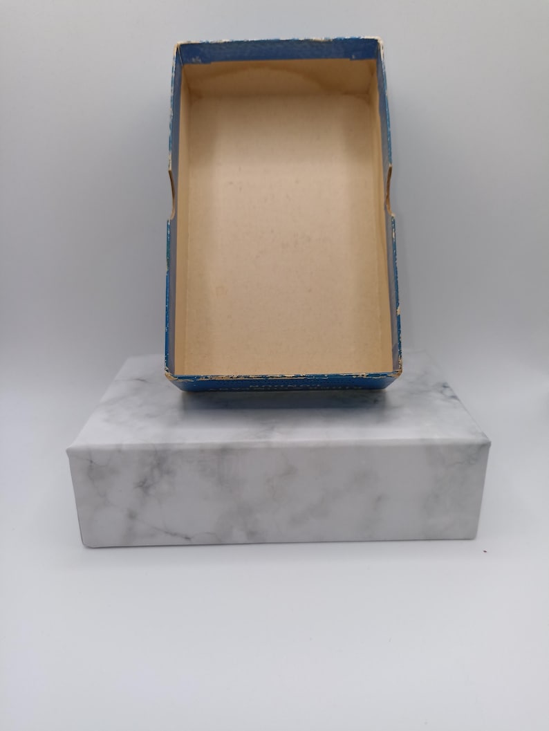 Puede incluir: Una caja rectangular abierta con borde azul e interior beige se encuentra sobre un bloque de m&aacute;rmol blanco. La caja parece ser vintage, con signos de desgaste. El bloque de m&aacute;rmol tiene un patr&oacute;n veteado gris.