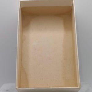 Puede incluir: Una caja rectangular vac&iacute;a con un interior beige claro y un exterior blanquecino. La caja tiene un dise&ntilde;o simple y limpio con una superficie ligeramente texturizada. La caja est&aacute; abierta, revelando su interior.