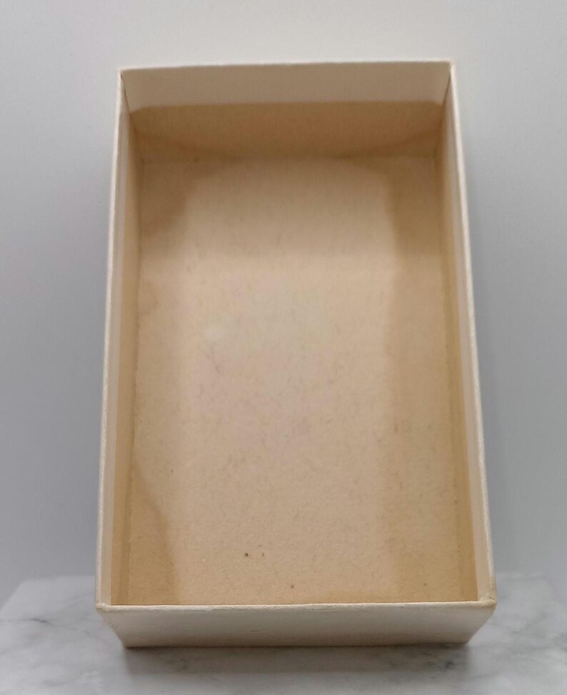Puede incluir: Una caja rectangular vac&iacute;a con un interior beige claro y un exterior blanquecino. La caja tiene un dise&ntilde;o simple y limpio con una superficie ligeramente texturizada. La caja est&aacute; abierta, revelando su interior.