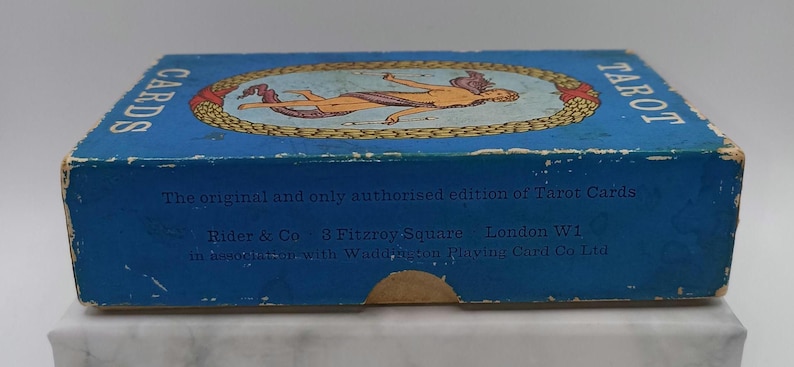 Puede incluir: Una caja de cartas de tarot azul vintage con las palabras "CARDS" y "TAROT". La caja presenta una ilustraci&oacute;n de una figura en un &oacute;valo dorado. El texto en la caja dice "The original and only authorised edition of Tarot Cards."