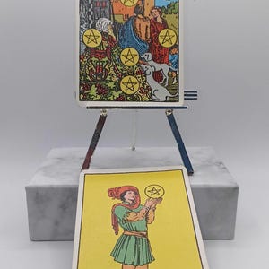 Puede incluir: Dos cartas de tarot, el Diez de Oros y la Sota de Oros, se exhiben en un peque&ntilde;o caballete. El Diez de Oros muestra una escena familiar con diez oros. La Sota de Oros muestra una figura sosteniendo un oro. Las cartas est&aacute;n sobre un bloque de m&aacute;rmol.