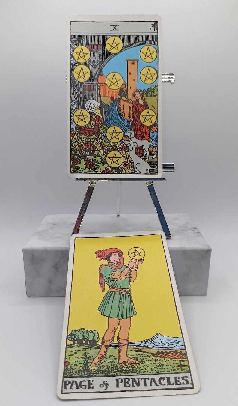 Puede incluir: Dos cartas de tarot, el Diez de Oros y la Sota de Oros, se exhiben en un peque&ntilde;o caballete. El Diez de Oros muestra una escena familiar con diez oros. La Sota de Oros muestra una figura sosteniendo un oro. Las cartas est&aacute;n sobre un bloque de m&aacute;rmol.