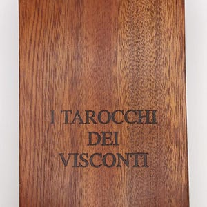 Puede incluir: Una caja de madera con el texto "I Tarocchi Dei Visconti" grabado en la parte frontal. La caja es de color marrón oscuro con vetas de madera visibles. El texto está en una fuente serif. La parte inferior de la caja tiene el texto "Accademia Carrara".