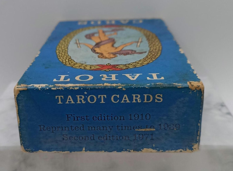 Puede incluir: Una caja de cartas del tarot azul vintage con la palabra "TAROT" en la parte superior. La caja presenta una ilustraci&oacute;n y el texto "TAROT CARDS, First edition 1910, Reprinted many times to 1989, Second edition 1971."