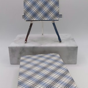 Puede incluir: Una baraja de cartas del tarot con un patr&oacute;n de cuadros azules, blancos y negros. Una carta se muestra en un caballete plateado en miniatura. Una pila de cartas descansa debajo sobre una superficie blanca. Las cartas son para la adivinaci&oacute;n y la adivinaci&oacute;n.