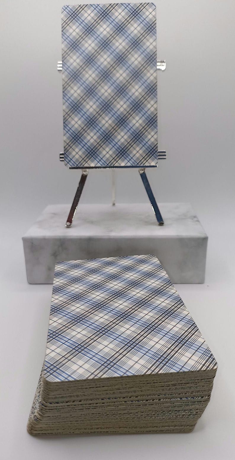 Puede incluir: Una baraja de cartas del tarot con un patr&oacute;n de cuadros azules, blancos y negros. Una carta se muestra en un caballete plateado en miniatura. Una pila de cartas descansa debajo sobre una superficie blanca. Las cartas son para la adivinaci&oacute;n y la adivinaci&oacute;n.