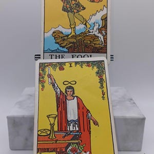 Puede incluir: Se muestran dos cartas del tarot. La carta superior es "El Loco", que representa una figura con un bast&oacute;n y una bolsa. La carta inferior es "El Mago", que muestra a una persona con t&uacute;nicas rojas sosteniendo una varita. Ambas cartas tienen fondos amarillos.