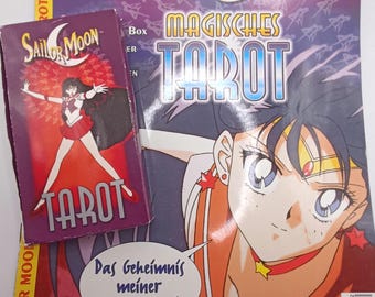 Könnte beinhalten: Eine Sailor Moon Magisches Tarot Kartendeckschachtel mit einer Figur in rot-weißem Outfit, platziert auf einem passenden Magazin mit dem Titel Sailor Moon Magisches Tarot, einer großen Illustration und dem Text Das Geheimnis meiner Tarotkarten.