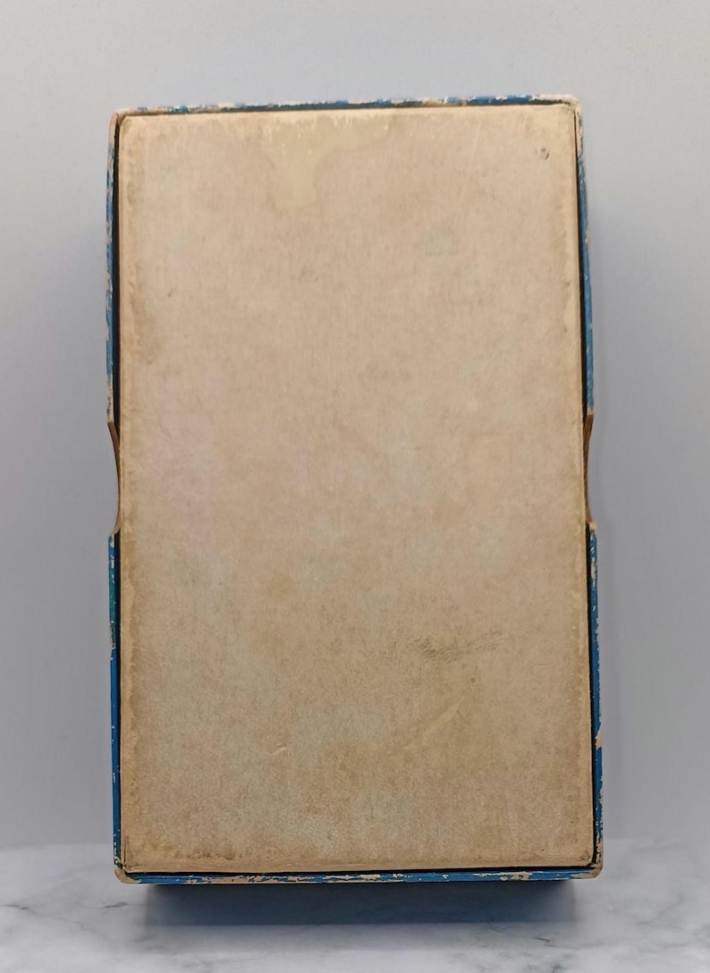 Puede incluir: Una caja rectangular vintage con tapa de color beige claro y ribete azul. La caja muestra signos de antig&uuml;edad, con decoloraci&oacute;n y desgaste en la superficie de papel. Los bordes de la caja est&aacute;n ligeramente desgastados.