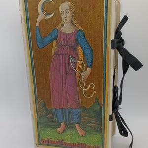 Op de afbeelding: Een vintage boek met een gedetailleerde illustratie op de omslag. Het kunstwerk toont een vrouw die een halve maan en een wit lint vasthoudt. De vrouw draagt een roze jurk en blauwe mouwen. Het boek is gebonden met een zwart lint.