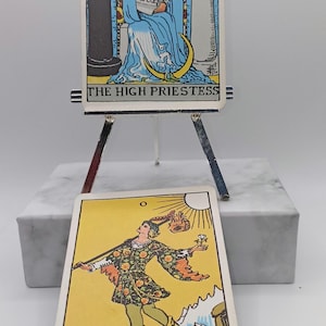 Puede incluir: Dos cartas de tarot, "La Suma Sacerdotisa" y "El Loco", se exhiben en un peque&ntilde;o caballete. La carta de la Suma Sacerdotisa presenta a una mujer con t&uacute;nicas azules. La carta del Loco muestra a una persona con un perro. Las cartas est&aacute;n sobre una base de m&aacute;rmol blanco.