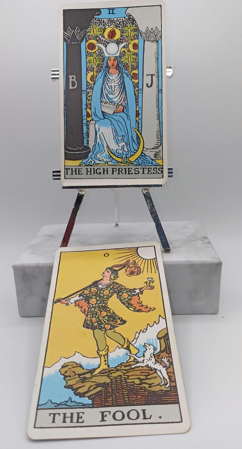 Puede incluir: Dos cartas de tarot, "La Suma Sacerdotisa" y "El Loco", se exhiben en un peque&ntilde;o caballete. La carta de la Suma Sacerdotisa presenta a una mujer con t&uacute;nicas azules. La carta del Loco muestra a una persona con un perro. Las cartas est&aacute;n sobre una base de m&aacute;rmol blanco.