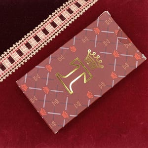 Puede incluir: Caja rectangular burdeos con un patrón repetido de coronas y la letra "I". La caja presenta una letra "I" dorada con una corona encima. Un ribete de encaje blanco y rojo es visible en la esquina superior izquierda.