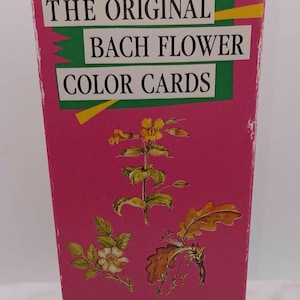 Op de afbeelding: Een rechthoekige roze doos met het opschrift THE ORIGINAL BACH FLOWER COLOR CARDS in zwarte letters op witte rechthoeken. De doos bevat illustraties van gele en witte bloemen met groene bladeren en bruine eikenbladeren. Ingrid S. Kraaz/Wulfing von Rohr.