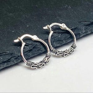 May include: A pair of small, silver hoop earrings. Each earring features a decorative, patterned design along the lower half of the hoop. The earrings are displayed on a dark, textured surface.