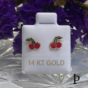 May include: A pair of cherry-shaped stud earrings. Each earring features two red cherries with green leaves and gold-colored outlines. The earrings are displayed on a white textured surface with the text "14 KT GOLD".