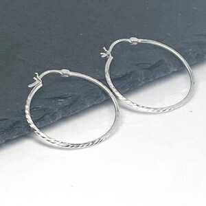 May include: Two silver hoop earrings with a textured, diamond-cut design. The earrings are displayed on a dark gray surface against a white background. The hoops have a classic, minimalist style.