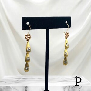 May include: A pair of gold drop earrings with three teardrop-shaped charms. Each charm has a small star design in the center. The earrings are hanging from a black display stand.