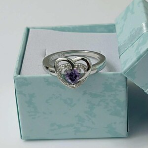 May include: A silver ring with a heart-shaped purple gemstone center, surrounded by smaller clear stones. The ring is set in a light blue gift box. The band is marked with "925".