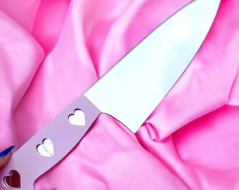 May include: A pink and silver kitchen knife with a heart-shaped handle. The knife has three cut-out hearts on the handle.