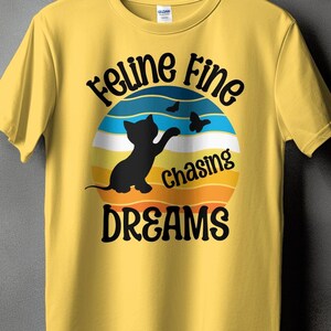 Puede incluir: Camiseta amarilla con un gráfico retro de un gato negro persiguiendo mariposas. El gráfico dice "Feline Fine Chasing Dreams".
