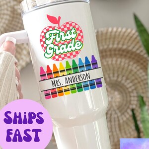 Könnte beinhalten: Ein weißer Edelstahlbecher mit einem roten und weißen Apfeldesign, auf dem "First Grade" steht, und einer Reihe bunter Stifte mit dem Namen "Mrs. Anderson".