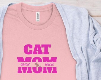 Peut inclure: T-shirt rose avec le texte "CAT MOM" en lettres roses. Les textes "GRACIE" et "ROCCO" sont écrits en lettres plus petites sous le texte principal, avec une silhouette de chat entre les deux noms.