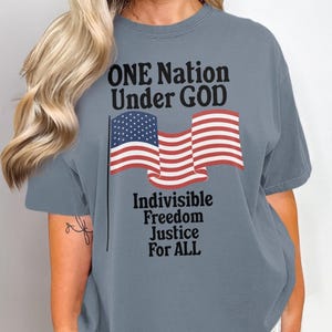 May include: A gray t-shirt with the text "ONE Nation Under GOD" and an American flag. Below the flag is the text "Indivisible Freedom Justice For ALL".