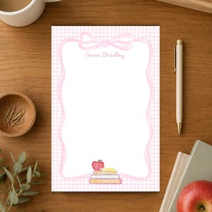 May include: A rectangular notepad featuring a light pink gingham background, a white central area framed by a pink ribbon border, and a watercolor illustration of a red apple resting on a stack of books. The name Anna Bradley is printed at the top.