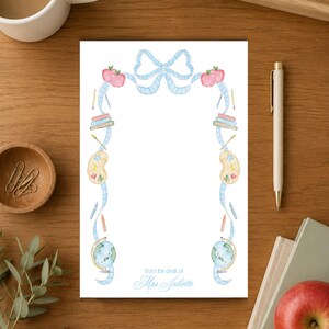 May include: A white rectangular notepad featuring a light blue gingham ribbon border decorated with watercolor illustrations of red apples, stacked books, paint palettes, pencils, and globes, with the text from the desk of Mrs. Juliette.
