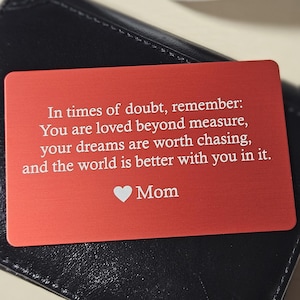 May include: A red metal card with the text "In times of doubt, remember: You are loved beyond measure, your dreams are worth chasing, and the world is better with you in it. Mom" is on top of a black leather wallet.